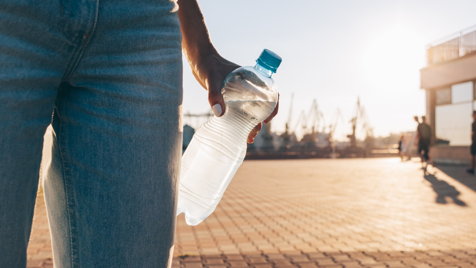 Weibliche Figur in Bluejeans mit Wasserflasche in der Hand steht draußen bei sonnigem Wetter